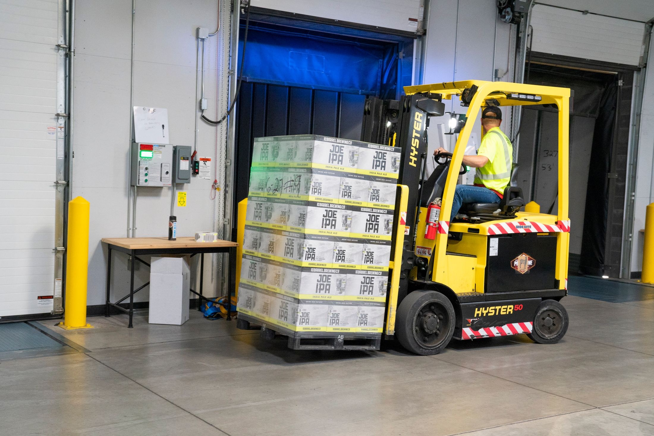 A forklift operator moving a wrapped pallet of goods at a warehouse loading dock.