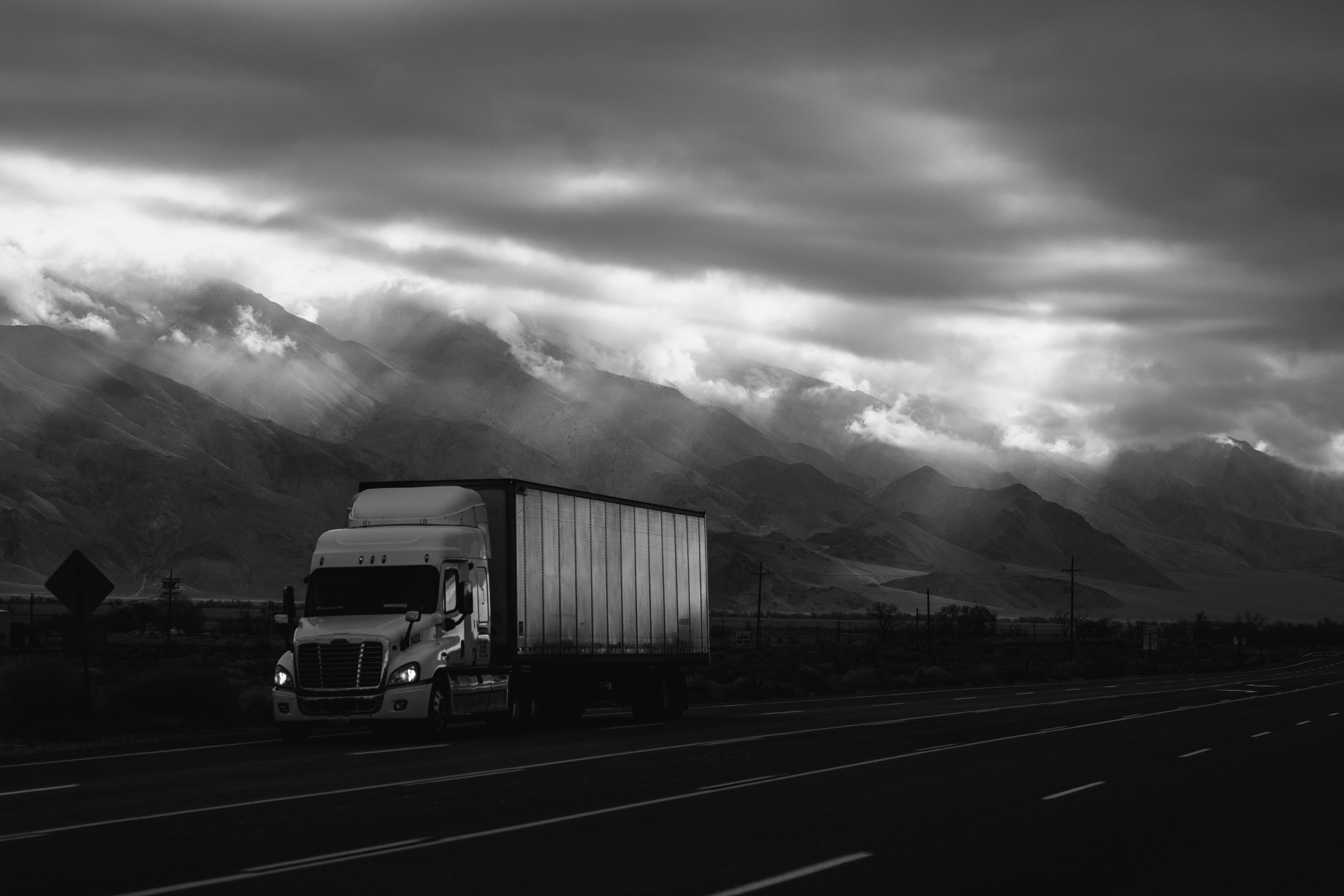 Black-and-white photograph of a conventional-cab tractor trailer traveling past mountains.